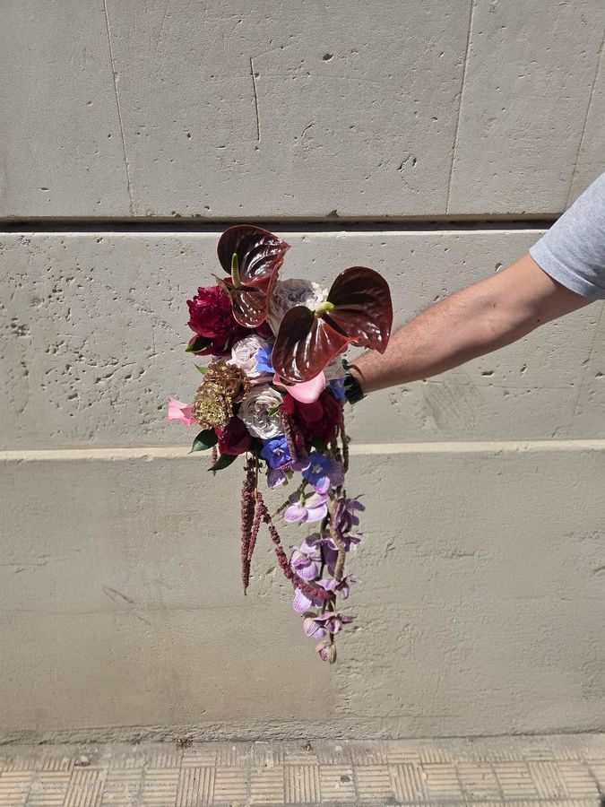 Bouquet a cascata con anthurium bordeaux, rose, peonia, delphinium e orchidee lilla su fondo muro in cemento