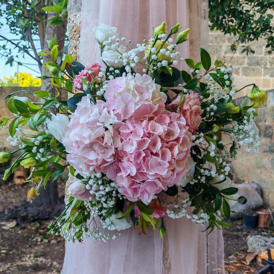 Bouquet tondo di ortensie rosa, lisianthus, garofani e gypsophila con fogliame verde, tenuto davanti a un abito lungo di tulle rosa cipria