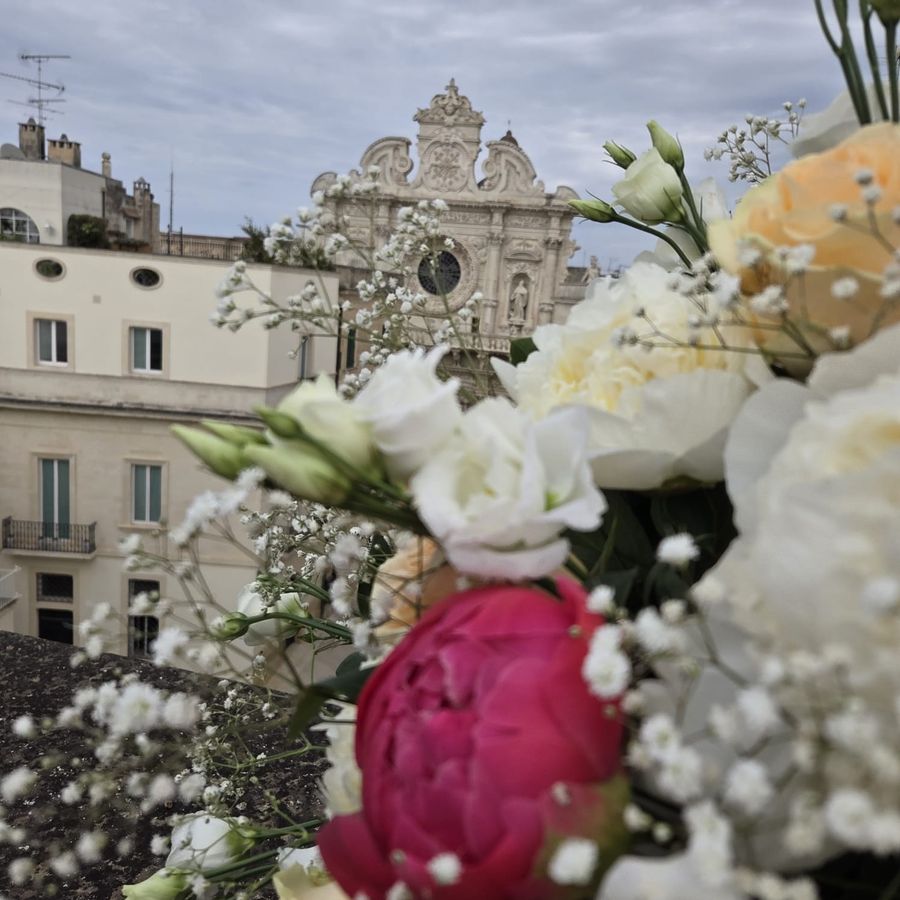 Mazzo di rose crema, peonia fucsia, lisianthus bianchi e nebbiolina appoggiato su un parapetto con la Basilica di Santa Croce di Lecce sullo sfondo