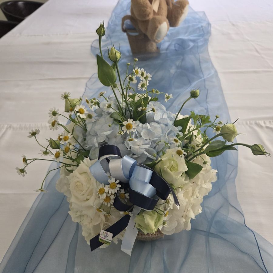 Composizione da battesimo con ortensie bianche e azzurre, rose e camomilla, fiocchi blu e celesti su runner in organza