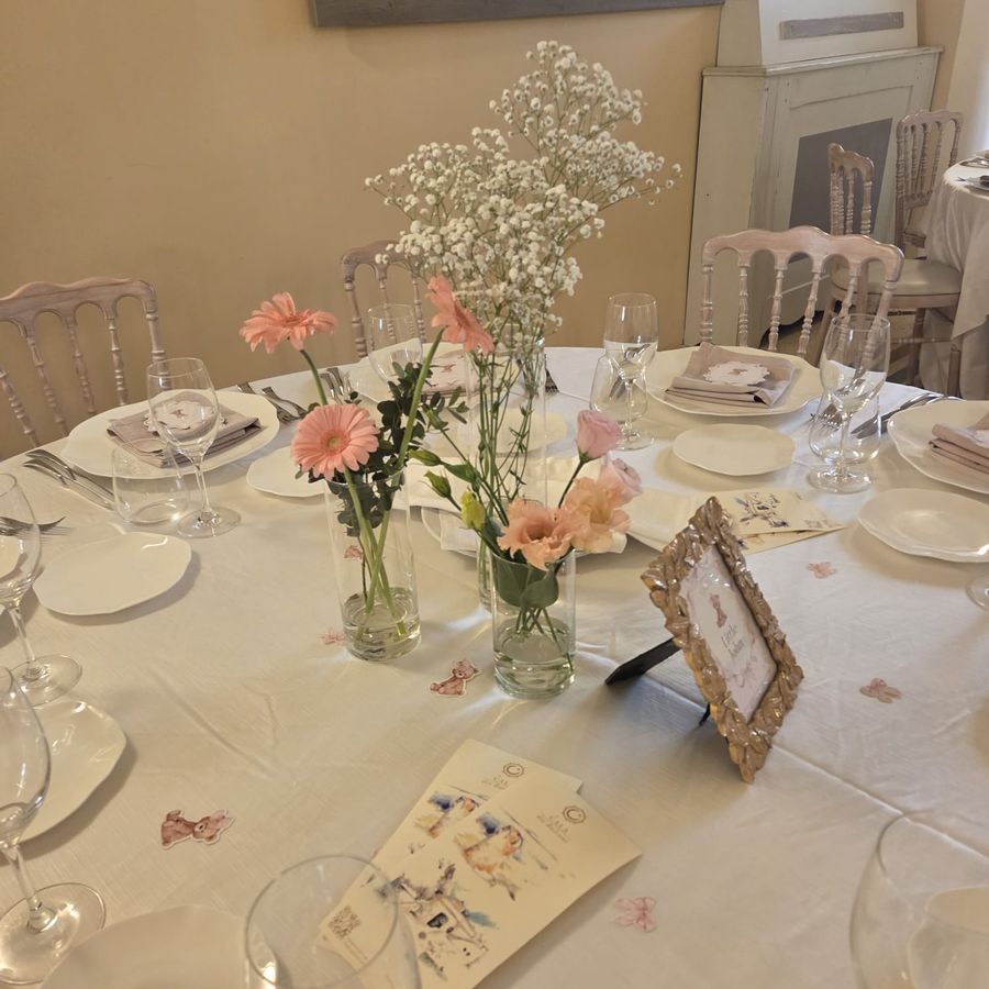 Tavolo allestito per battesimo con vasetti cilindrici di gerbere rosa, lisianthus e nebbiolina, cornice segnatavolo e coriandoli a forma di orsetto