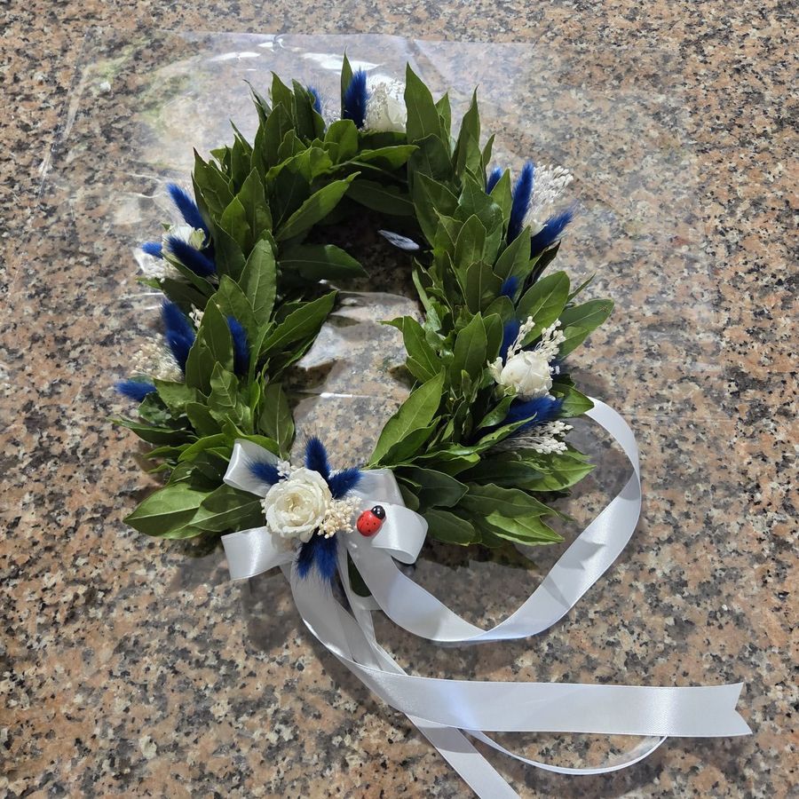 Corona d'alloro da laurea decorata con lagurus blu, roselline bianche stabilizzate, gypsophila, coccinella portafortuna e nastro di raso bianco