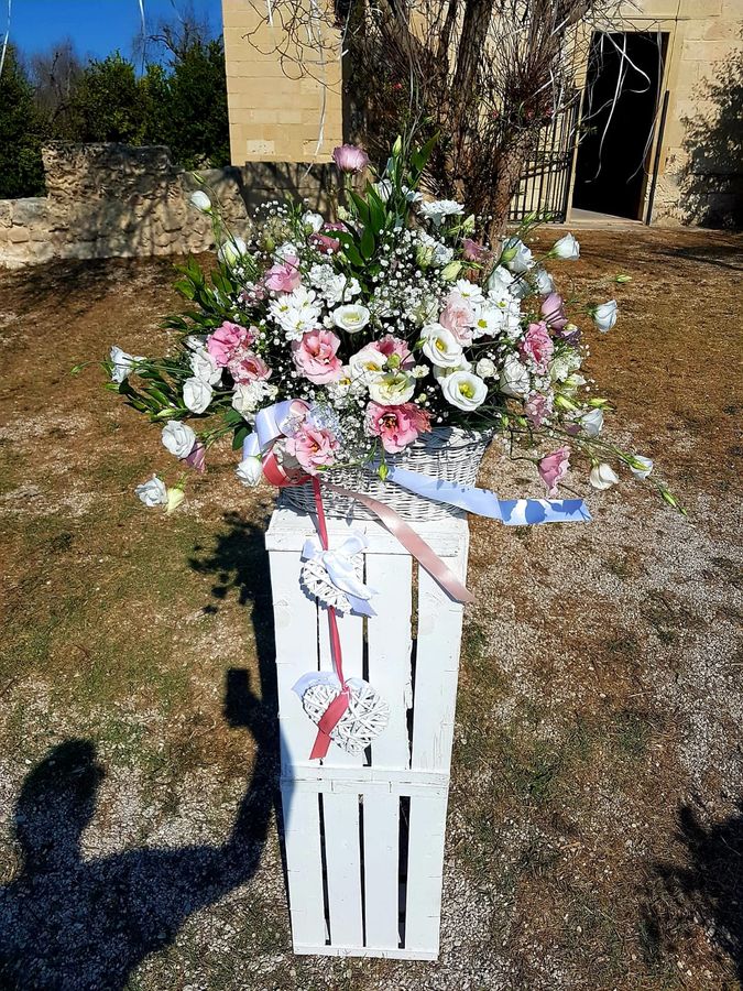 Cesto in vimini bianco con lisianthus rosa e bianchi, margherite e gypsophila, appoggiato su una cassetta di legno bianca decorata con cuori in vimini e nastri