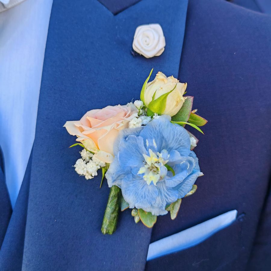 Boutonniere con rosa cipria, delphinium azzurro e gypsophila su abito da sposo blu con pochette