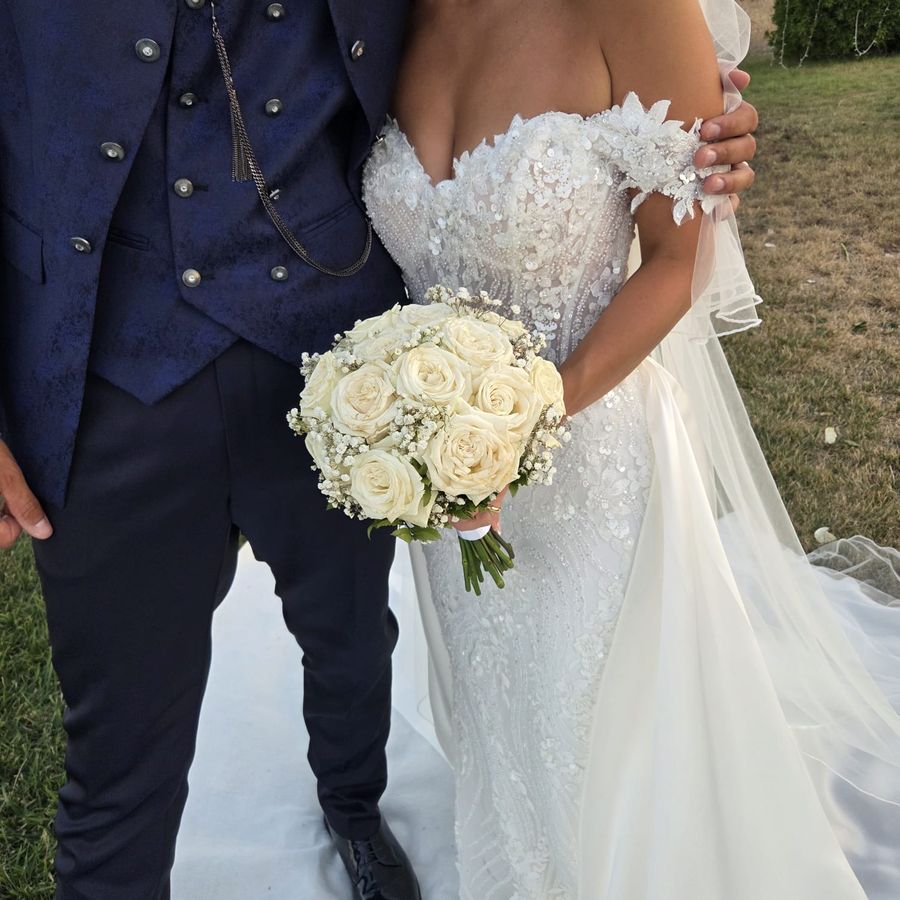 Bouquet da sposa tondo di rose bianche e gypsophila tra le mani della sposa accanto allo sposo