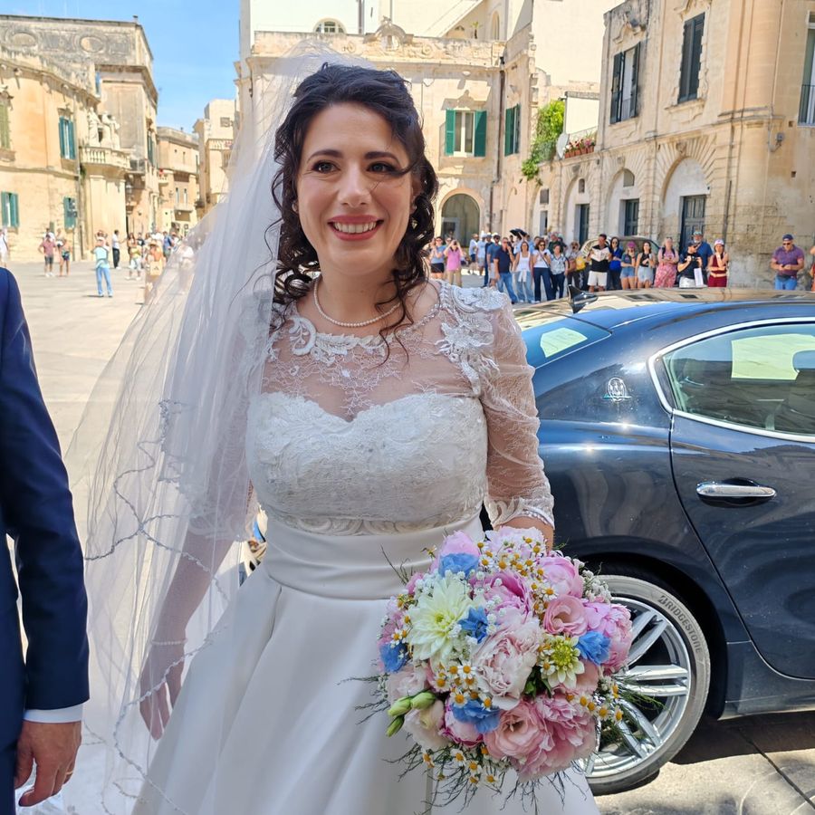 Sposa sorridente in abito di pizzo bianco con velo, bouquet tondo di rose rosa, dalie bianche e delphinium azzurro, accanto all'auto degli sposi nel centro storico