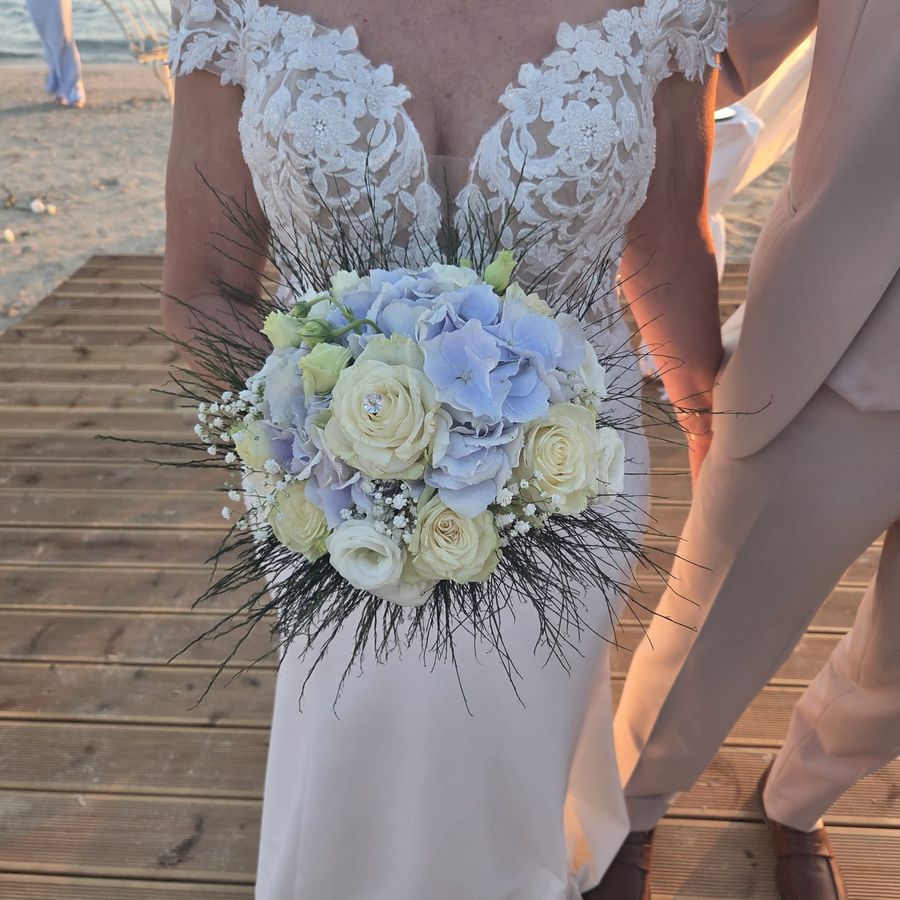 Sposa in abito di pizzo che tiene un bouquet tondo di ortensie azzurre, rose bianche e gypsophila su passerella al mare