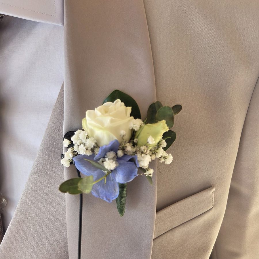 Boutonniere da sposo con rosa bianca, delphinium azzurro ed eucalipto appuntata sul bavero di un abito beige
