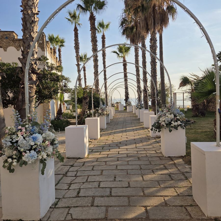 Viale cerimoniale con archi bianchi e composizioni di ortensie azzurre, rose e lisianthus bianchi su colonne, con vista mare tra le palme