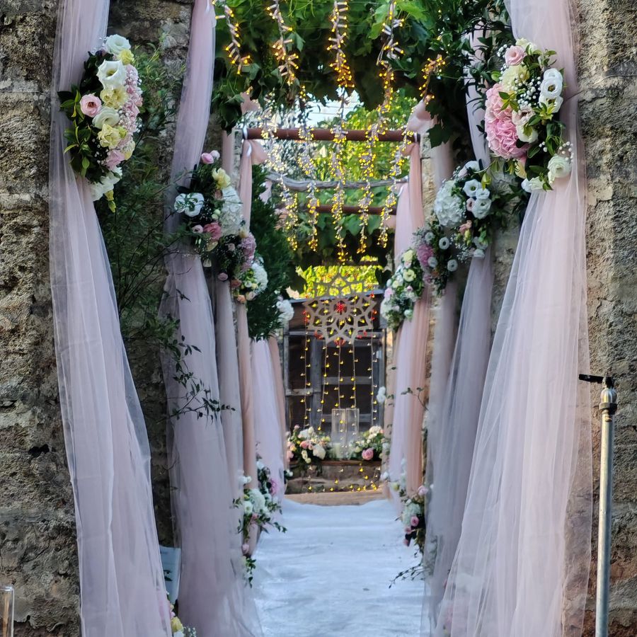 Corridoio di cerimonia in pietra addobbato con drappi di tulle rosa cipria, composizioni di ortensie e rose e catene di lucine