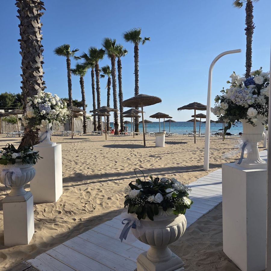 Allestimento cerimonia in spiaggia con vasi in pietra su colonne bianche e composizioni di rose e ortensie azzurre lungo la passerella