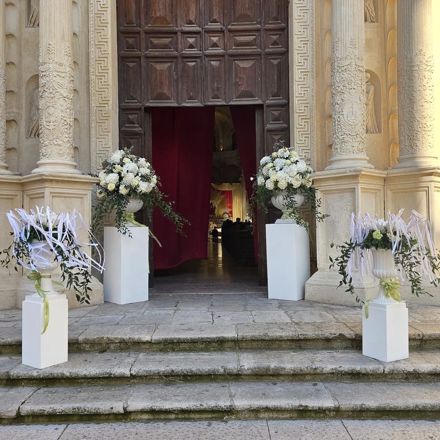 Ingresso della chiesa addobbato con quattro coppe bianche su colonnine, rose e ortensie bianche e nastri di raso