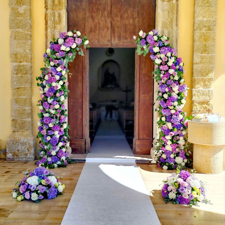 Arco floreale di ortensie lilla, rose bianche ed eucalipto sul portale di una chiesa con passatoia nuziale