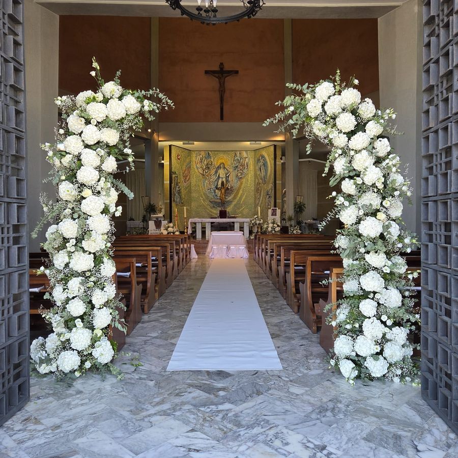 Arco floreale bianco di ortensie, rose e gypsophila all'ingresso della navata, con tappeto bianco verso l'altare