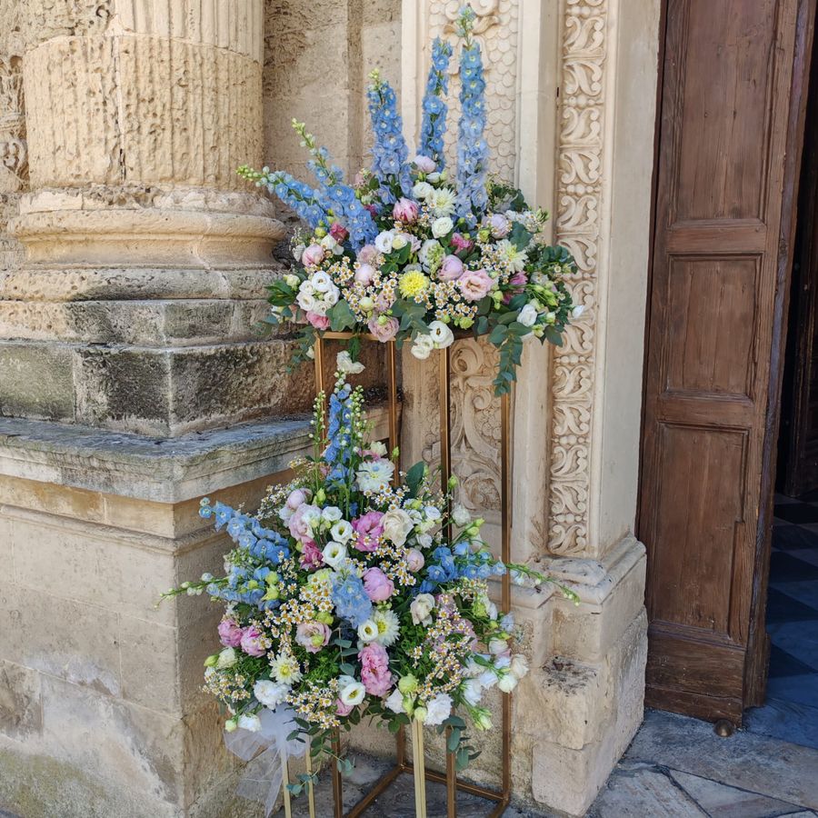 Due composizioni floreali su alzate dorate con delphinium azzurro, peonie rosa, lisianthus e margheritine, all'ingresso di una chiesa in pietra leccese