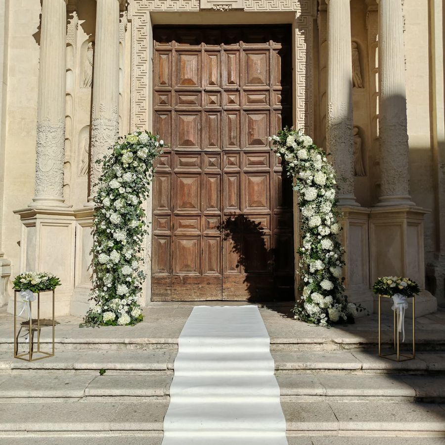Arco floreale asimmetrico di ortensie e rose bianche con eucalipto sul portale di una chiesa, con tappeto bianco e due alzate dorate con bouquet