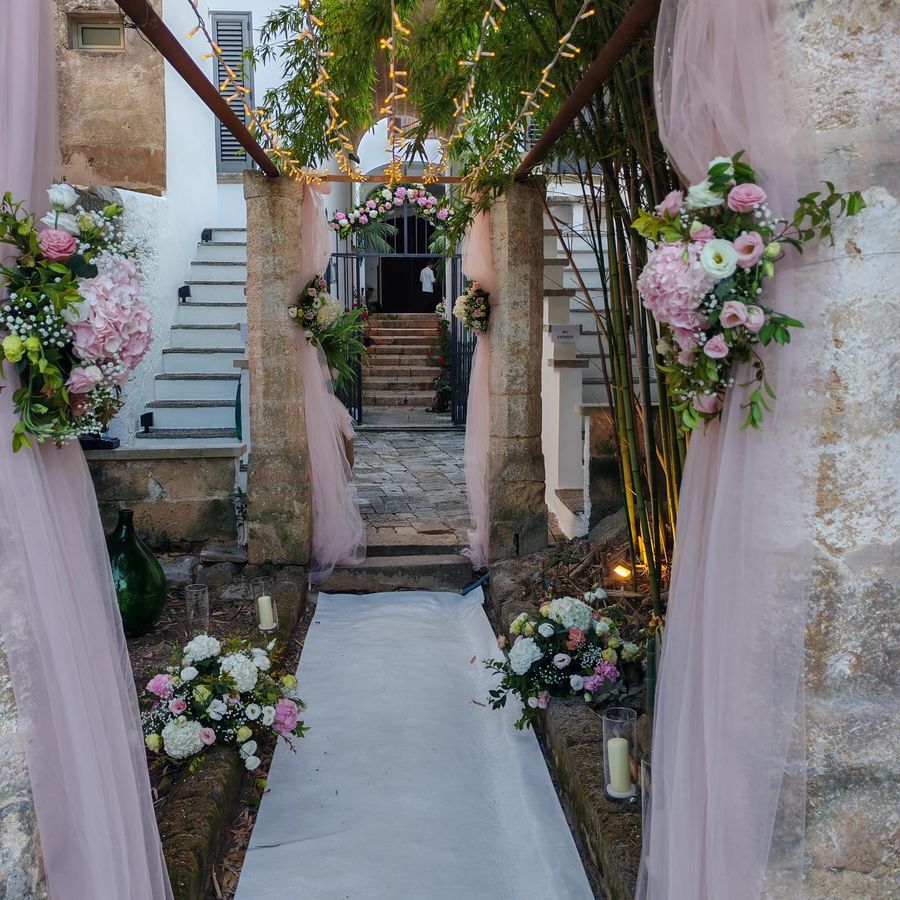 Ingresso di masseria addobbato per una cerimonia con drappi di tulle rosa, composizioni di ortensie e rose, tappeto bianco, candele e luci calde