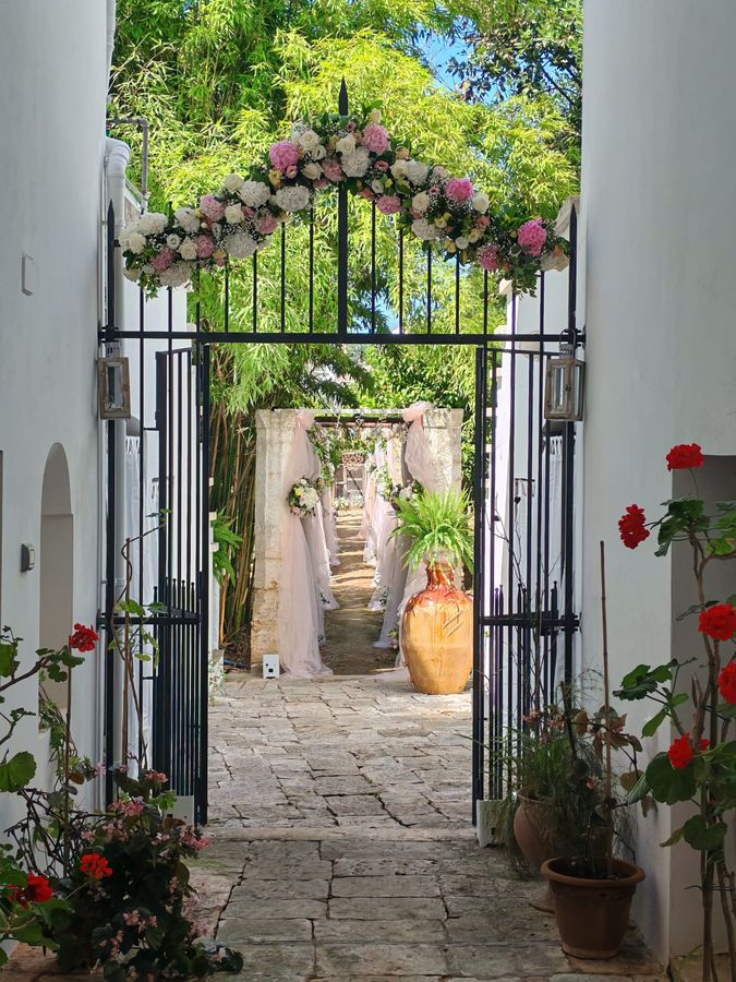 Cancello in ferro battuto sormontato da un arco floreale di rose bianche, ortensie rosa e gypsophila, con vialetto in pietra addobbato di tulle rosa e bouquet laterali