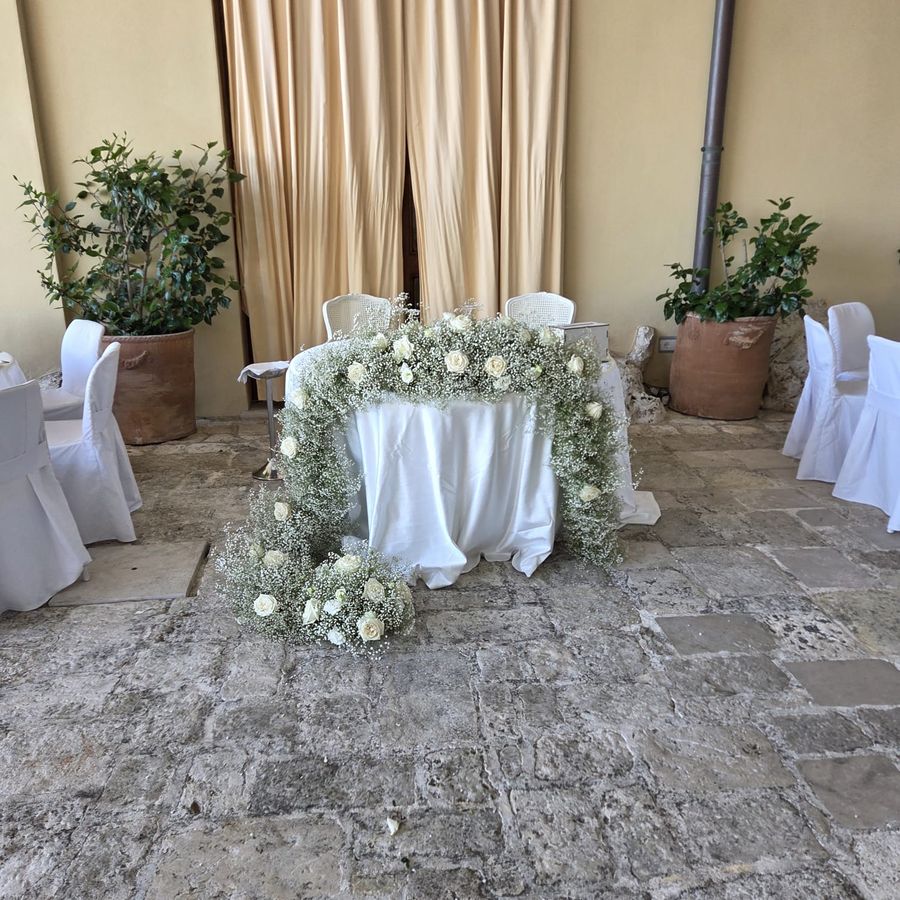 Tavolo degli sposi avvolto da un arco di gypsophila e rose bianche, in una corte in pietra con sedie vestite di bianco