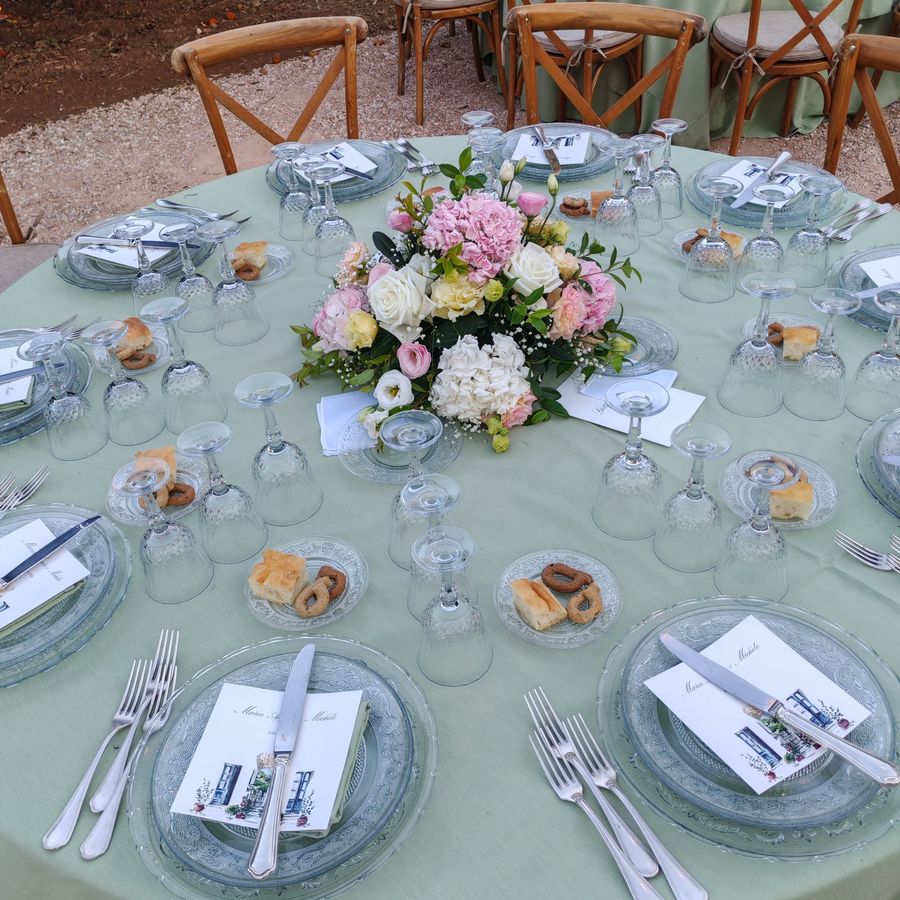 Tavolo rotondo di ricevimento nuziale con tovaglia verde salvia, piatti in vetro e centrotavola di ortensie rosa, rose bianche e lisianthus