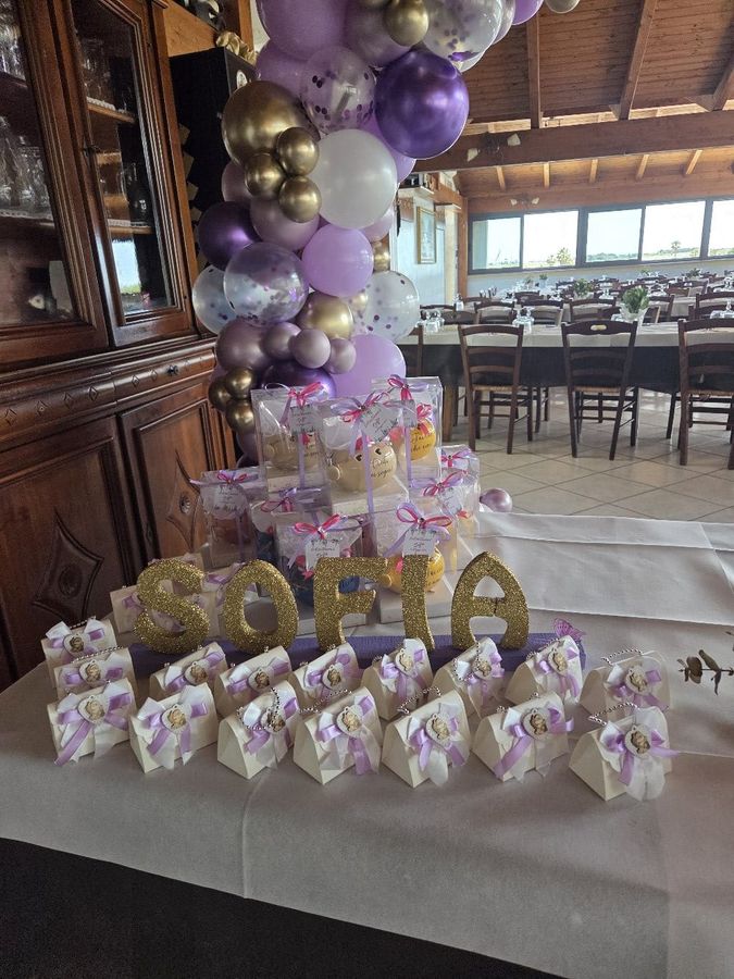 Tavolo delle bomboniere con sacchetti portaconfetti bianchi e nastri lilla, scatoline trasparenti e nome in lettere dorate glitter